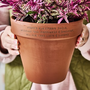 Può includere: Un vaso di fiori in terracotta marrone pieno di piante rosa e verdi. Il vaso ha un messaggio scritto a mano: "HAPPY BIRTHAY JULIA, WISHING YOU HAPPINESS & SUNSHINE, ALL OUR LOVE LUCY & SIMONX."