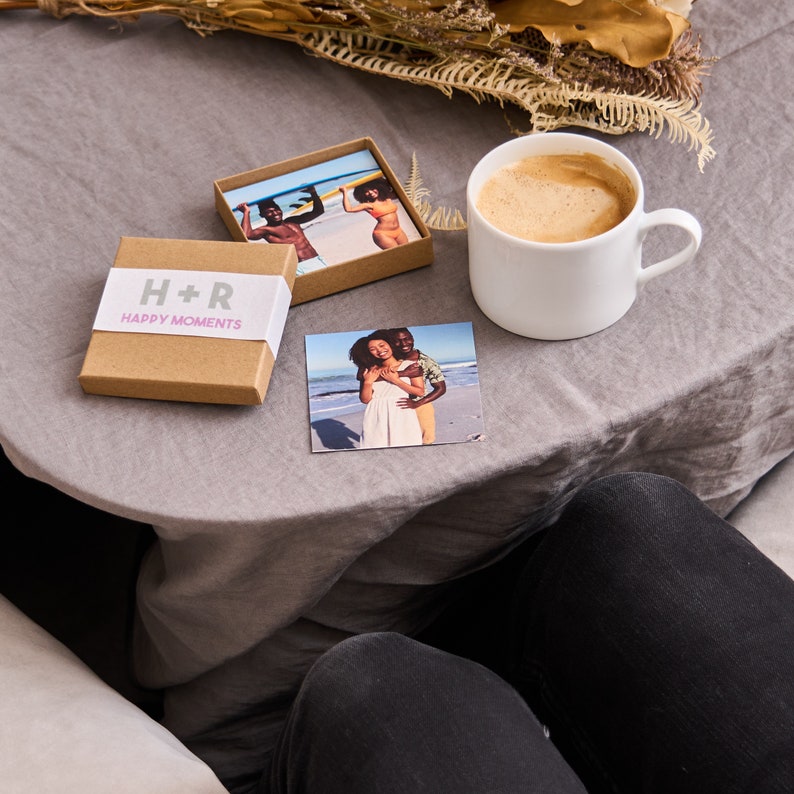 Puede incluir: Una caja marr&oacute;n con el texto "H+R Happy Moments" en la parte delantera. La caja est&aacute; sobre un mantel gris con una foto de una pareja en la playa y una taza de caf&eacute;.
