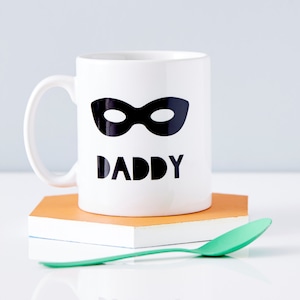 Peut inclure: Une tasse en c&eacute;ramique blanche avec un masque noir sur les yeux et le mot "DADDY" imprim&eacute; dessus. La tasse est pos&eacute;e sur un dessous-de-tasse brun et blanc.