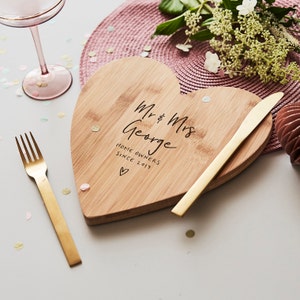 Puede incluir: Tabla de servir de madera en forma de corazón con la inscripción "Mr & Mrs George HOME OWNERS SINCE 2017" y un símbolo de corazón. Un cuchillo y un tenedor dorados están colocados cerca de la tabla. La tabla descansa sobre un mantel individual rosa.