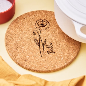 May include: A round cork trivet with a black line drawing of a poppy flower and the name "Bella" engraved on it.