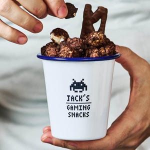 Pode incluir: Uma caneca de esmalte branco com borda azul, cheia de pipocas cobertas com chocolate e um palito de chocolate. A caneca tem um gráfico alienígena pixelado e o texto "JACK'S GAMING SNACKS". Uma mão está adicionando mais pipoca.