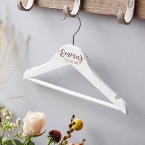 Può includere: Un appendiabiti in legno bianco con il nome "Emma's" e le parole "Christening Gown" incise in oro. L'appendiabiti è appeso a un gancio a muro. Lo sfondo è grigio chiaro. I fiori sono in primo piano.