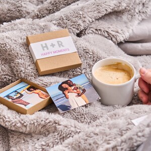 Puede incluir: Una caja marr&oacute;n con el texto "H+R Happy Moments" en la parte delantera. La caja est&aacute; abierta y contiene peque&ntilde;as tarjetas de fotos con im&aacute;genes de una pareja. Las tarjetas de fotos est&aacute;n sobre una superficie gris y peluda con una taza de caf&eacute; blanca.