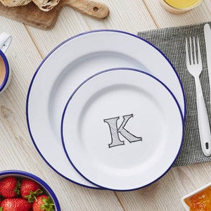 May include: Two white enamel plates with blue rims. The smaller plate has a black and white illustration of the letter K in the centre.
