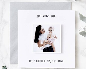 May include: A white greeting card with a photograph of a mother and baby. The card reads "BEST MUMMY EVER" and "HAPPY MOTHER'S DAY, LOVE EMMA" in black text. A gray envelope and decorative ribbon are also visible.