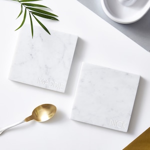 May include: Two white marble coasters with the names "Maddie" and "Nick" engraved on them. The coasters are square and have a light grey veining.