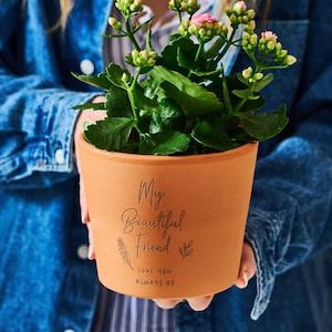 Puede incluir: Una maceta de color terracota con las palabras "My Beautiful Friend" y "Love You Always XX" impresas. La maceta contiene una planta verde con flores rosas. La maceta es sostenida por una persona con una chaqueta vaquera.