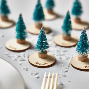 Puede incluir: Una colección de pequeños árboles de Navidad verde azulado sobre bases de madera, algunos con nombres grabados. Los árboles tienen un aspecto flocado y están colocados sobre una superficie blanca con nieve artificial, cerca de un plato y un tenedor.