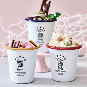 Pode incluir: Três canecas de esmalte branco com texto preto e gráficos de estrelas, cheias de lanches. Uma caneca tem uma borda azul e é preenchida com chocolates. Outra tem uma borda amarela e contém doces rosa. A terceira tem uma borda vermelha e é preenchida com pipocas.