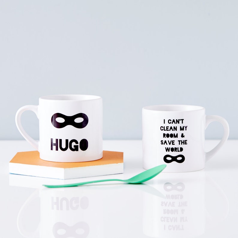 Peut inclure: Deux tasses en c&eacute;ramique blanche avec du texte noir et un motif de masque noir. Une tasse dit "HUGO" et l'autre tasse dit "Je ne peux pas nettoyer ma chambre et sauver le monde".