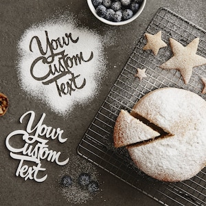 Pode incluir: Um bolo redondo polvilhado com açúcar de confeiteiro, com uma fatia removida, está sobre uma grelha. Biscoitos em forma de estrela e uma tigela de mirtilos estão por perto. As palavras "Your Custom Text" são escritas com farinha.