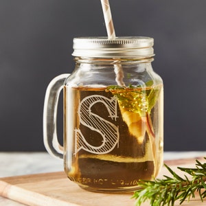 May include: A clear glass mason jar with a handle, a straw, and a lid. The jar has an etched letter "S" on the front and is filled with a light brown liquid. The jar is sitting on a wooden surface.