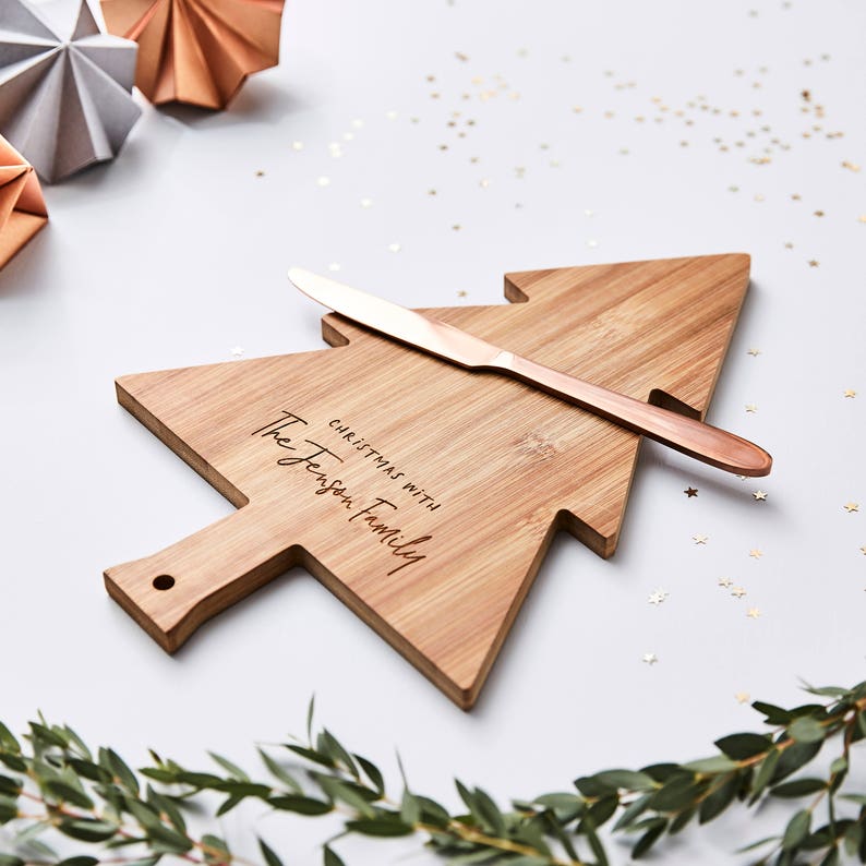 Pode incluir: Uma t&aacute;bua de servir de bambu em forma de &aacute;rvore de Natal com o texto gravado "CHRISTMAS WITH The Jenson Family" e uma faca de manteiga de cor cobre met&aacute;lico pousada horizontalmente sobre o centro da superf&iacute;cie de madeira.
