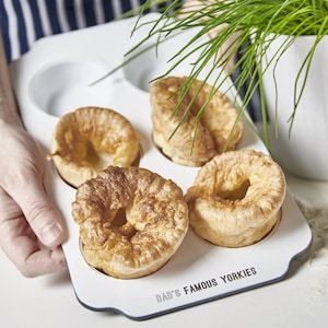 Op de afbeelding: Een witte bakplaat met vier goudbruine Yorkshire puddings. De bakplaat heeft een geschulpte rand en de tekst "DAD'S FAMOUS YORKIES". Verse bieslook staat op de achtergrond in een witte pot.