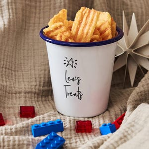 Pode incluir: Uma caneca de esmalte branco com borda azul, cheia de salgadinhos dourados e canelados. A caneca tem o texto "Leo's Treats" e um desenho de estrela. Blocos de construção de brinquedo coloridos estão espalhados.