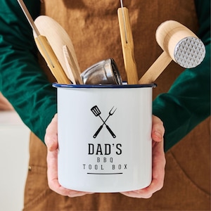 May include: White enamelware utensil holder with a blue rim. The holder has a black graphic of crossed grilling tools and the text "Dad's BBQ Toolbox".