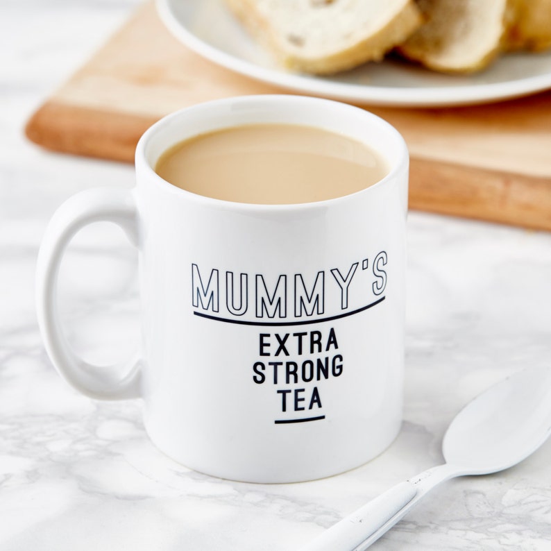 Puede incluir: Taza de cer&aacute;mica blanca con texto negro que dice "Mummy's Extra Strong Tea". La taza est&aacute; llena de un l&iacute;quido marr&oacute;n claro y est&aacute; sobre una superficie de m&aacute;rmol blanco.