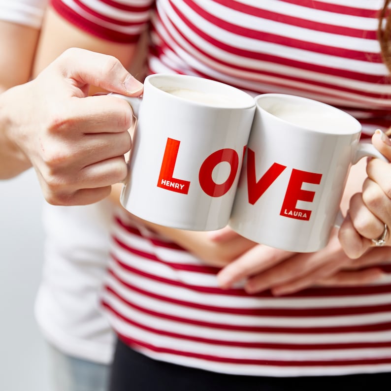 May include: Two white ceramic mugs with the word "LOVE" in red, and the names "Henry" and "Laura" below. The mugs are held by two people, one wearing a red and white striped top.