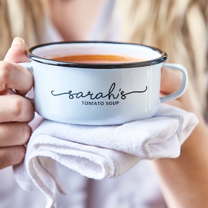 Può includere: Tazza per zuppa in smalto bianco con bordo e manico neri. La tazza ha la scritta "sarah's TOMATO SOUP" stampata sul davanti.