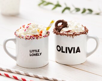 Op de afbeelding: Twee witte mokken met slagroom en toppings. De ene mok heeft sprankelingen en zegt "Little Lovely" en de andere heeft chocoladeschilfers en zegt "Olivia".