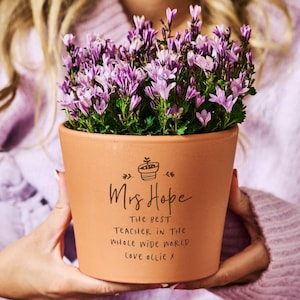 Puede incluir: Una maceta de terracota llena de flores moradas. La maceta tiene el texto "Mrs Hope, The Best Teacher in the Whole Wide World, Love Ollie X". La maceta es sostenida por una persona que lleva un suéter morado.