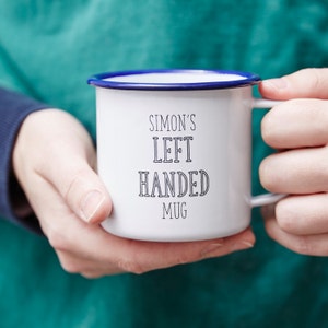 Może przedstawiać: Biały emaliowany kubek z niebieską obw&oacute;dką i uchwytem, trzymany przez osobę. Kubek ma napis "SIMON'S LEFT HANDED MUG" czarnymi literami. Kubek jest przeznaczony dla os&oacute;b leworęcznych.