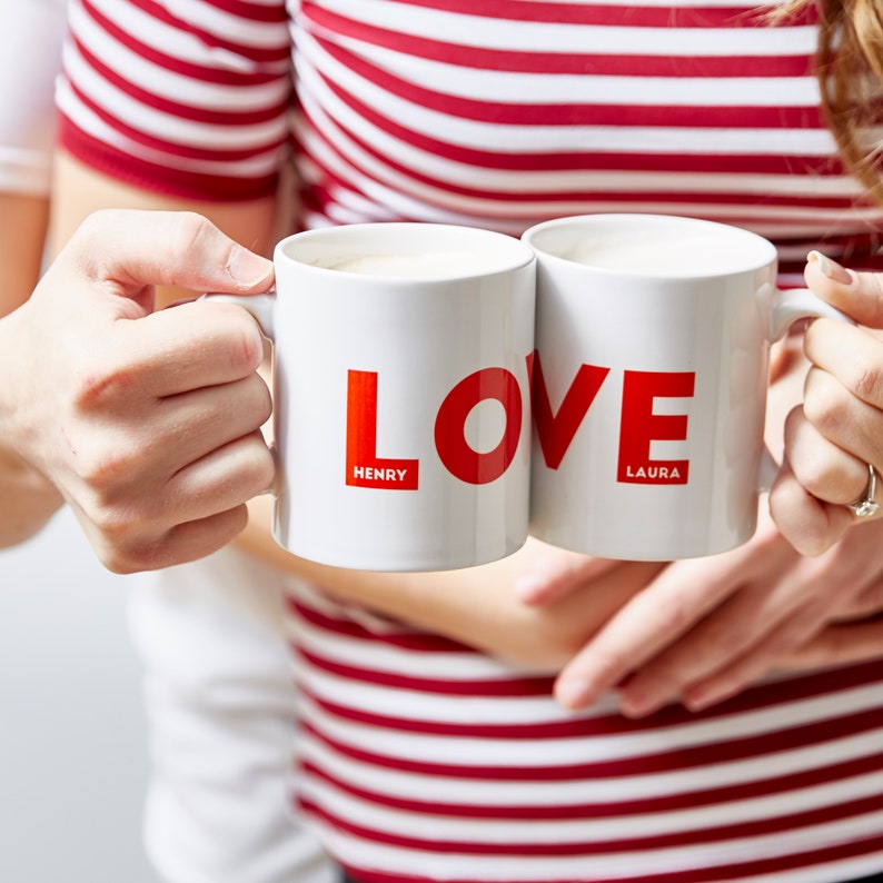 May include: Two white ceramic mugs with the word "LOVE" in red block letters. The names "HENRY" and "LAURA" are printed in red below the letters. The mugs are held by two people, one wearing a red and white striped top.