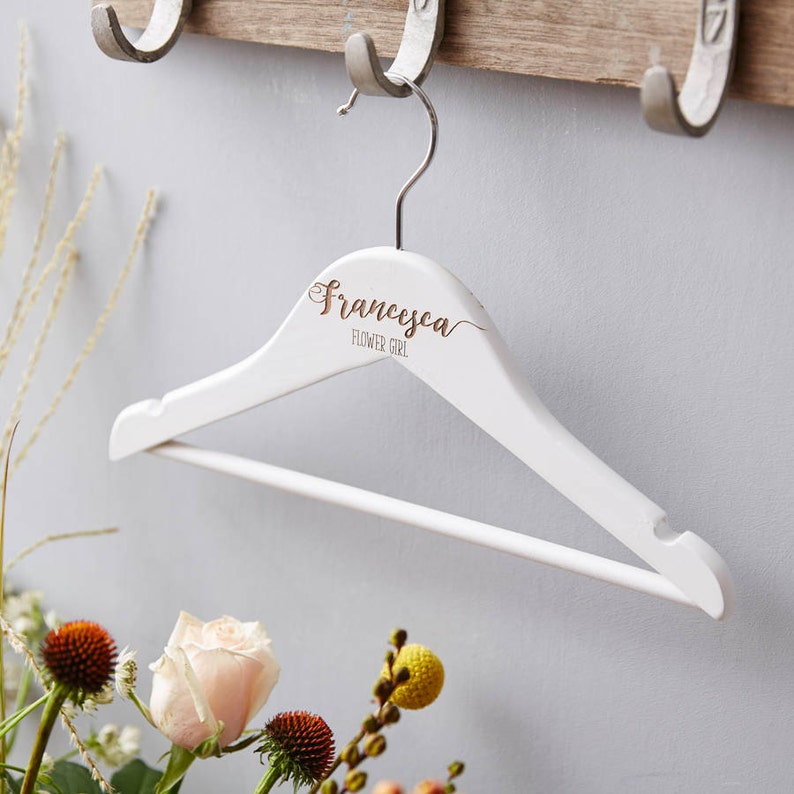 May include: White wooden hanger with the name "Francesca" and the words "Flower Girl" written in gold lettering.