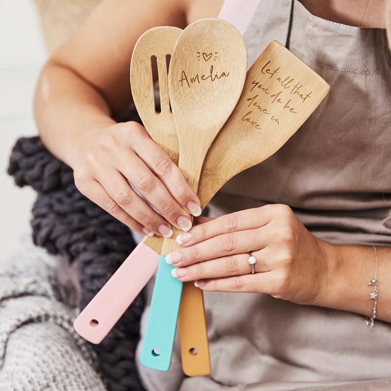 May include: Three wooden kitchen utensils with colorful painted handles. The utensils are a slotted spoon, a large spoon, and a spatula. The spoons are engraved with the text "Amelia" and "Let all that you do be done in love".