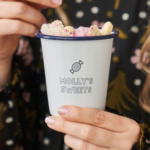 May include: White enamel mug with a blue rim, filled with colourful sweets. The mug has a black and white logo that says "Molly's Sweets" and a sweet graphic.