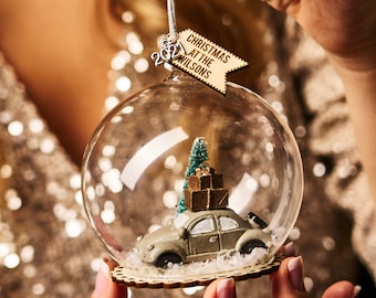 May include: A glass Christmas ornament with a miniature car carrying a small Christmas tree and presents inside. The ornament has a wooden tag that reads "Christmas at the Wilsons 2021".