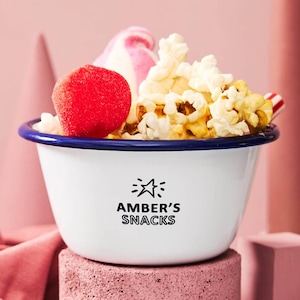 Pode incluir: Uma tigela de esmalte branco com borda azul, cheia de pipocas, um doce vermelho e um doce rosa e branco em espiral. A tigela tem o texto "AMBER'S SNACKS" impresso na frente. A tigela está em um bloco rosa.