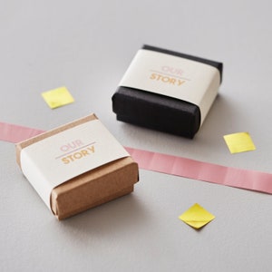 May include: Two small gift boxes, one brown and one black, are displayed on a gray surface. Each box is wrapped with a cream-colored band printed with the words "OUR STORY" in pink and gold. Pink ribbon and yellow confetti add color.