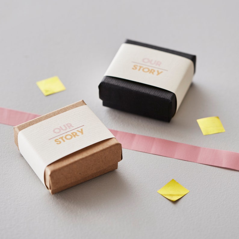 May include: Two small gift boxes, one brown and one black, are displayed on a gray surface. Each box is wrapped with a cream-colored band printed with the words "OUR STORY" in pink and gold. Pink ribbon and yellow confetti add color.