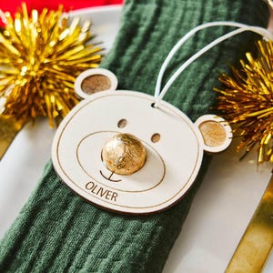 Könnte beinhalten: Ein hölzerner Bären-Namensschildhalter mit dem eingravierten Namen "OLIVER". Der Bär hat eine goldverpackte Süßigkeiten-Nase und liegt auf einer grünen Serviette. Goldener Lametta und ein goldenes Utensil sind im Hintergrund.