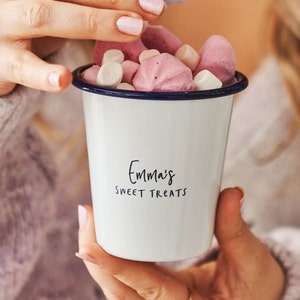 Pode incluir: Caneca de esmalte branco com borda azul cheia de marshmallows rosa e brancos. A caneca tem o texto "Emma's Sweet Treats" impresso na frente.