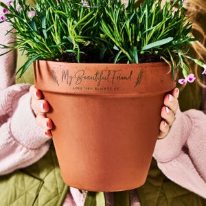 May include: A brown terracotta plant pot filled with green foliage and small pink flowers. The pot is inscribed with "My Beautiful Friend" and "Love You Always XX" in black script. The pot is held by a person.