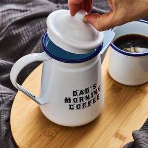 Pode incluir: Bule de café de esmalte branco com borda azul e uma inscrição preta que diz "Dad's Morning Coffee". A bule tem uma tampa branca e uma alça. A bule está sobre uma superfície de madeira.