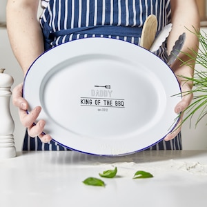 Op de afbeelding: Witte ovale serveerschaal met een blauwe rand. De schaal heeft de zwarte tekst "DADDY KING OF THE BBQ EST. 2012". Een houten lepel en tang zijn zichtbaar achter de schaal.