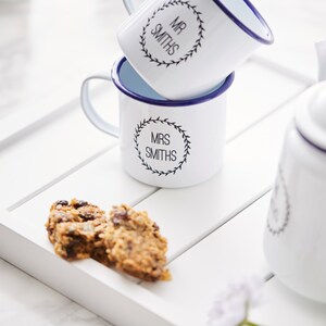 Puede incluir: Dos tazas de esmalte blanco con bordes azules y letras negras. Las tazas est&aacute;n apiladas una encima de la otra. La taza superior dice "Mr. Smiths" en un dise&ntilde;o de corona. La taza inferior dice "Mrs. Smiths" en un dise&ntilde;o de corona. Las tazas est&aacute;n en una bandeja de madera blanca con dos galletas de avena.