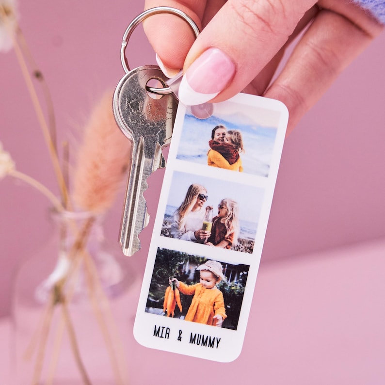Close-up of a Personalised Photo Strip Keyring featuring three custom photo slots and the names 'MIA & MUMMY'; a Custom Photo Gift Keyring and Personalized Photo Memory Keychain attached to a silver keyring, held against a soft pink background.