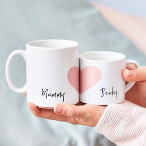以下が含まれることがあります： ピンクのハートが付いた白いセラミック製のマグカップ2つ。大きなマグカップには「Mummy」と書いてあり、小さなマグカップには「Becky」と書いてあります。