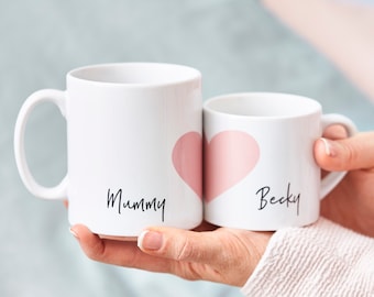 Puede incluir: Dos tazas de cerámica blanca con corazones rosas. La taza más grande dice "Mummy" y la taza más pequeña dice "Becky".