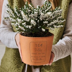 May include: A terracotta plant pot containing a white flowering heather plant. The pot features a black printed rectangular border with the text: Daddy THANK YOU FOR HELPING US GROW love Ella & Mia. The plant has dense green foliage and white flowers.