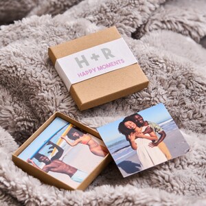 Puede incluir: Una caja marr&oacute;n con una etiqueta blanca que dice "H+R Happy Moments". La caja est&aacute; abierta y contiene dos fotos peque&ntilde;as. Una foto muestra una pareja en una playa, y la otra foto muestra a una persona sosteniendo una tabla de surf.
