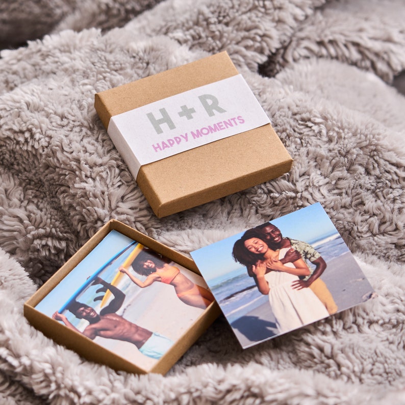 Puede incluir: Una caja marr&oacute;n con una etiqueta blanca que dice "H+R Happy Moments". La caja est&aacute; abierta y contiene dos fotos peque&ntilde;as. Una foto muestra una pareja en una playa, y la otra foto muestra a una persona sosteniendo una tabla de surf.