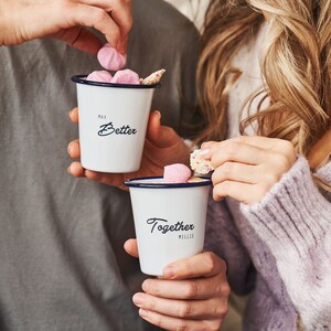 Pode incluir: Duas canecas de esmalte branco com bordas azuis. A caneca à esquerda diz "Max Better" e a caneca à direita diz "Together Millie".