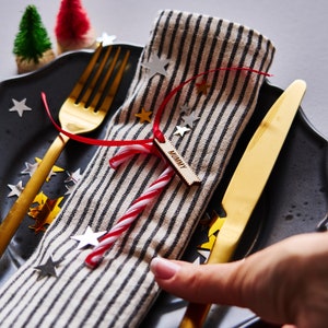 Puede incluir: Decoración de mesa festiva con plato negro, cubiertos dorados y servilleta a rayas. Un bastón de caramelo y una etiqueta de madera con la palabra "MUMMY" están atados a la servilleta con una cinta roja. Estrellas y árboles en miniatura.