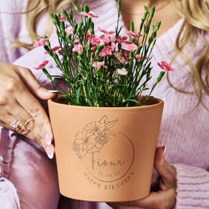 May include: A terracotta flower pot containing pink dianthus flowers. The pot features a circular floral line art design with the name Fiona, the date 12.09.15, and the text Happy Birthday engraved on the front surface of the clay material.